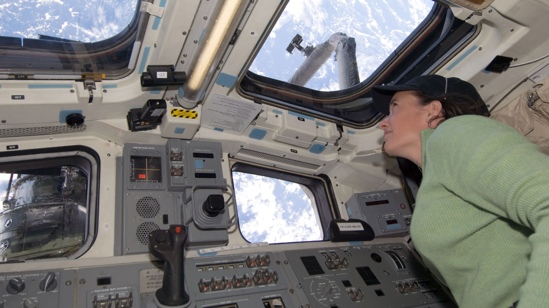 A woman wearing a headset and a green long sleeve shirt looks out of the windows on the International Space Station aimed at Earth