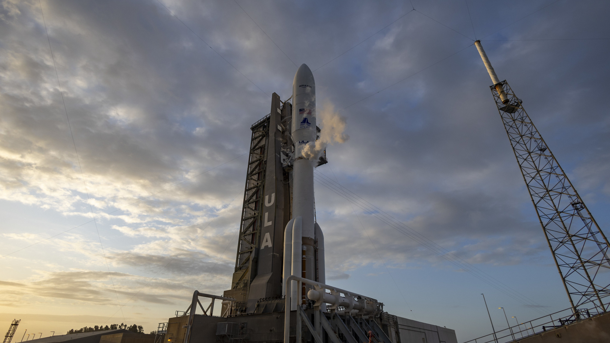 A white Atlas V rocket with strap on boosters vents gas during fueling for Amazon's Kuiper 1 satellite launch.