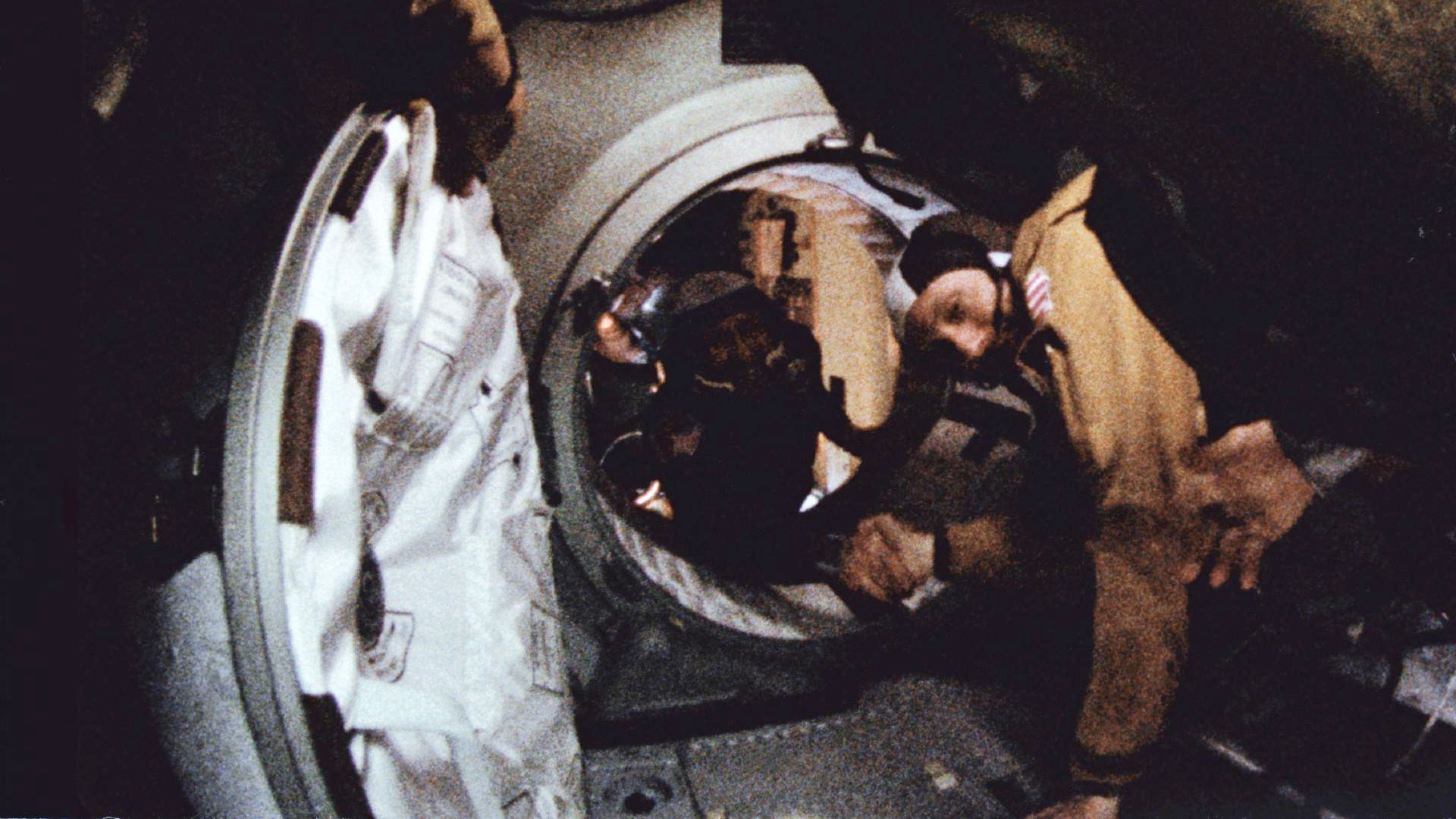 two men floating in space reach out and shake hands across the open threshold of a spacecraft hatchway