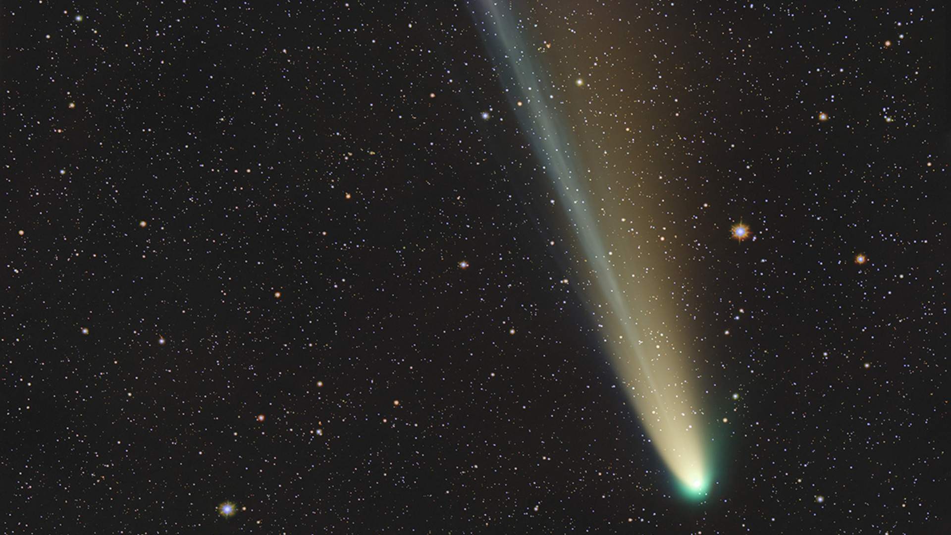 A bright comet is pictured against a black starry sky with its bright central coma visible to the lower left of the image from which a bright tail extends to the upper left.